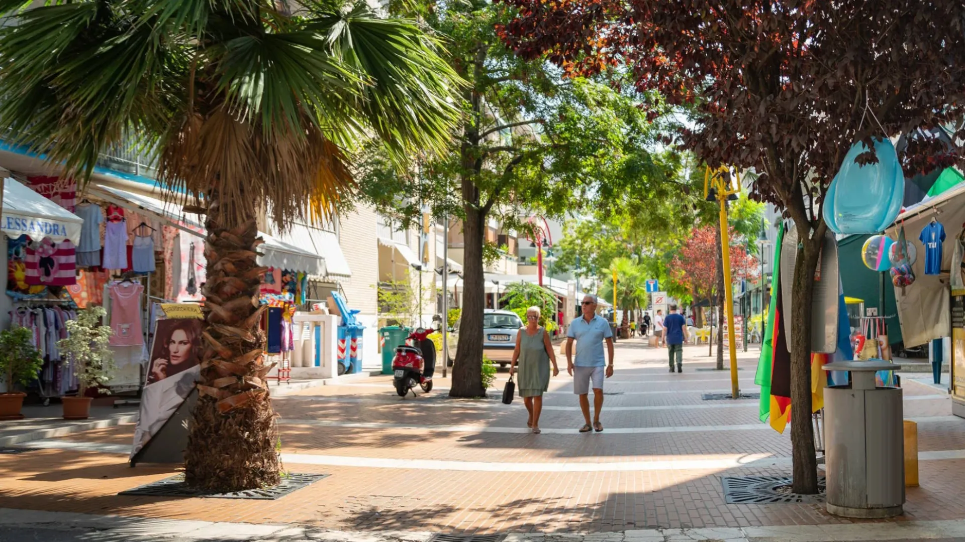 Strada pedonale con negozi, palmizi e persone che passeggiano in una giornata soleggiata.